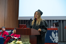 A person wearing a black academic gown and a black mortarboard speaks at a lectern with two microphones. In the background, people in red academic gowns and mortarboards are seated.