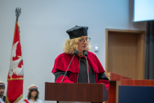 A woman in an academic gown and mortarboard speaks at the lectern. Behind her is a banner featuring a white eagle on a red background.