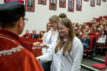Participants approach a university representative wearing an academic gown one by one during the ceremony. The audience seated in red chairs is visible in the background.