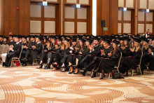 A large group of people in black academic gowns and mortarboards sit in rows and applaud during an academic ceremony.