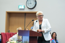 A person wearing a white blazer stands at a lectern with two microphones during a ceremony in an auditorium. Event participants are seated in the background, with a clock mounted above the door.