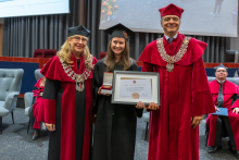 Three people pose during the ceremony. A person in a black academic gown and mortarboard holds a framed diploma and an open box containing a medal. The other two people are wearing red academic robes and ceremonial chains