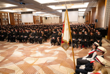 A view of a large hall filled with participants in black academic gowns and mortarboards; a ceremonial banner is visible in the foreground.