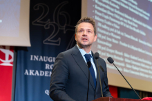 A person wearing a suit delivers a speech at the lectern, with the academic year inauguration stage in the background.