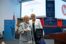 Two people pose for a photo in an auditorium. One person holds a glass award, while the other holds an open presentation box and a navy blue bag. In the background, a screen displays the text “Matriculation of students of the WUM Faculty of Health Sciences.