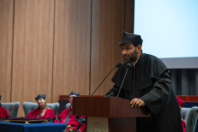 A person wearing a black academic gown, a black mortarboard and glasses speaks at a lectern with two microphones. In the background, people in red academic gowns are seated, and a white floral arrangement is in the foreground.