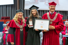 Three people pose during the ceremony. A person in a black academic gown and mortarboard holds a framed diploma and an open box containing a medal. The other two people are wearing red academic robes and ceremonial chains.