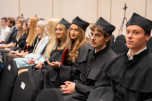 Participants in black academic gowns and mortarboards sit in a row during the ceremony; some are holding red folders.