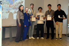 Six people pose for a photo after the competition. The participants are holding certificates and prizes, with a presentation about the event displayed in the background.