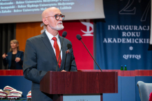 A person wearing a suit delivers a speech at the lectern, with the academic year inauguration stage in the background.