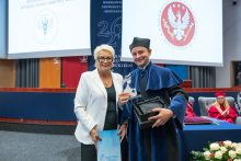 Two people stand next to each other in an auditorium. The person wearing a blue academic gown and black cap holds a glass award and an open presentation box, while the other person holds a blue folder. The Medical University of Warsaw emblem is displayed on a screen in the background.
