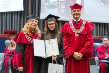 Three people pose during the ceremony. A person in a black academic gown and mortarboard holds a framed diploma and a bag. The other two people are wearing red academic robes and ceremonial chains.