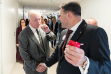 Two people shake hands in a hospital corridor. Event participants and a crew documenting the meeting can be seen in the background.