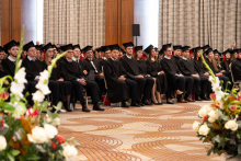 A large group of participants in black academic gowns and mortarboards sit in rows in a large hall during an academic ceremony; floral arrangements are visible in the foreground.