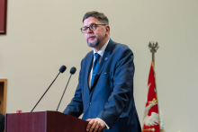 A person wearing a suit delivers a speech at the lectern, with the academic year inauguration stage in the background.
