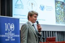 Prelegent w szarym garniturze przemawia do mikrofonu podczas prezentacji. Obok widoczny baner „100 lat Wydziału Farmaceutycznego WUM 1926–2026”.