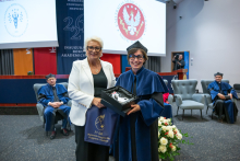 Two women stand next to each other during a ceremony in an auditorium. The woman wearing a blue academic gown and black cap holds an open box containing an award, while the woman in a white blazer holds a navy blue bag. People in academic gowns and screens displaying the symbols of the Medical University of Warsaw are in the background.