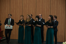 A vocal ensemble performs in the auditorium. Four people wearing black tops and long green skirts and four people wearing black suits hold open folders containing sheet music. Next to the ensemble stands a person holding a keyboard instrument.