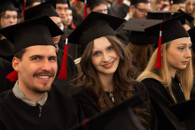 Three people in black academic gowns and mortarboards sit among the other participants in an academic ceremony.