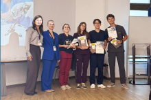 Six people pose for a photo after the competition. The participants are holding certificates and prizes, with a presentation about the event displayed in the background.