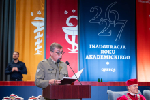 A person wearing a uniform delivers a speech at the lectern, with the academic year inauguration stage in the background.