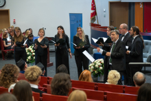 A vocal ensemble of four women and four men sings in front of an audience in an auditorium. The performers hold open folders with sheet music, while a man standing in front conducts the ensemble.