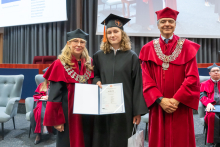 Three people pose during the ceremony. A person in a black academic gown and mortarboard holds a framed diploma and a bag. The other two people are wearing red academic robes and ceremonial chains.