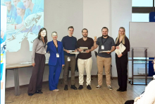 Six people pose for a photo after the competition. The participants are holding certificates and prizes, with a presentation about the event displayed in the background.