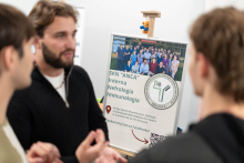 Three people talk at the SKN ANCA booth. In the background, a display board presents the group’s activities in internal medicine, nephrology, and immunology.