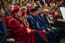 People wearing red and blue academic gowns and black and red caps sit in the front row of an auditorium. Some wear ceremonial chains over their gowns, while other ceremony participants are seated in the background.