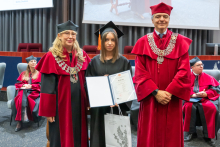 Three people pose during the ceremony. A person in a black academic gown and mortarboard holds a framed diploma and a bag. The other two people are wearing red academic robes and ceremonial chains.