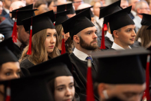 A close-up of people sitting next to one another in black academic gowns and mortarboards among the participants in an academic ceremony.