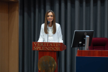 Kobieta z długimi brązowymi włosami ubrana w białą koszulę przemawia za mównicą z napisem „VERITATIS SPLENDOR” w auli uniwersyteckiej.