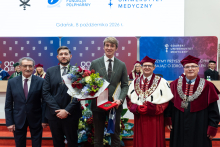Pięciu mężczyzn pozuje do wspólnego zdjęcia. Jeden z nich stojący w środku, profesor Piwowarski, trzyma bukiet kwiatów, medal w etui i dyplom. W tle widoczne są osoby siedzące przy stole prezydialnym oraz plansze z logo uczelni.