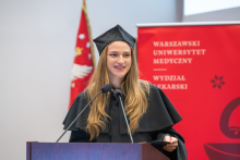 A young woman wearing a black academic gown stands at the lectern and delivers a speech. Part of the university banner and a burgundy backdrop featuring the faculty logo are visible in the background.