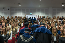 A large group of participants stand in rows in a crowded auditorium. People wearing blue and red academic gowns and black caps walk between the rows.
