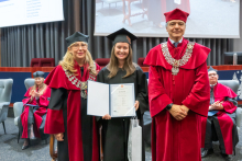 Three people pose during the ceremony. A person in a black academic gown and mortarboard holds a framed diploma and a bag. The other two people are wearing red academic robes and ceremonial chains.