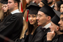People in black academic gowns and mortarboards sit in rows during an academic ceremony.