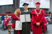 Three people pose during the ceremony. A person in a black academic gown and mortarboard holds a framed diploma and a bag. The other two people are wearing red academic robes and ceremonial chains.