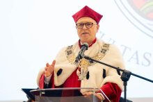 A person in red-and-white academic attire with a ceremonial chain speaks into a microphone at the lectern.