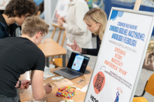 Participants talk at the booth of the Student Research Group in General and Vascular Surgery, Angiology and Phlebology. A laptop, instruments, and materials for practical training are displayed on the table.