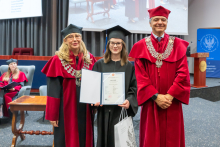 Three people pose during the ceremony. A person in a black academic gown and mortarboard holds a framed diploma and a bag. The other two people are wearing red academic robes and ceremonial chains.