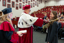 The Rector, wearing ceremonial attire, touches a female student’s shoulder with the ceremonial mace during the matriculation ceremony. A representative of the university authorities stands beside them holding an academic cap.