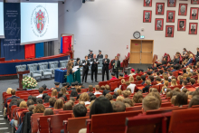 A vocal ensemble performs in front of an audience in a crowded auditorium. The performers hold open folders with sheet music, while the emblem of the Faculty of Medicine of the Medical University of Warsaw is displayed on the screen.