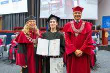 Three people pose during the ceremony. A person in a black academic gown and mortarboard holds a framed diploma and a bag. The other two people are wearing red academic robes and ceremonial chains.