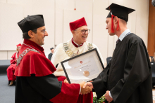 A person in a black academic gown and mortarboard receives a red folder and shakes hands with a person in ceremonial red-and-white academic attire.