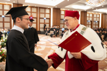 A person in a black academic gown and mortarboard receives a framed diploma during an academic ceremony and shakes hands with a person in red academic robes.