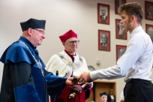 A student receives an academic cap from a representative of the university authorities wearing a blue academic gown. The Rector holding the ceremonial mace is visible in the background.