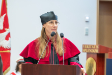 A woman in an academic gown and mortarboard speaks at the lectern. Behind her is a banner featuring a white eagle on a red background.