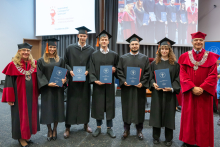 Seven people pose on stage during the ceremony. Five graduates in black academic gowns and mortarboards hold navy-blue folders, while two people in red academic robes stand on either side.